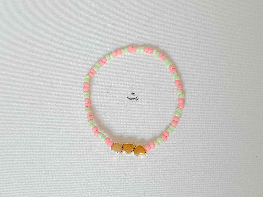 Flat lay of delicate pastel bracelet featuring soft pink and green beads with three golden heart accents in the center